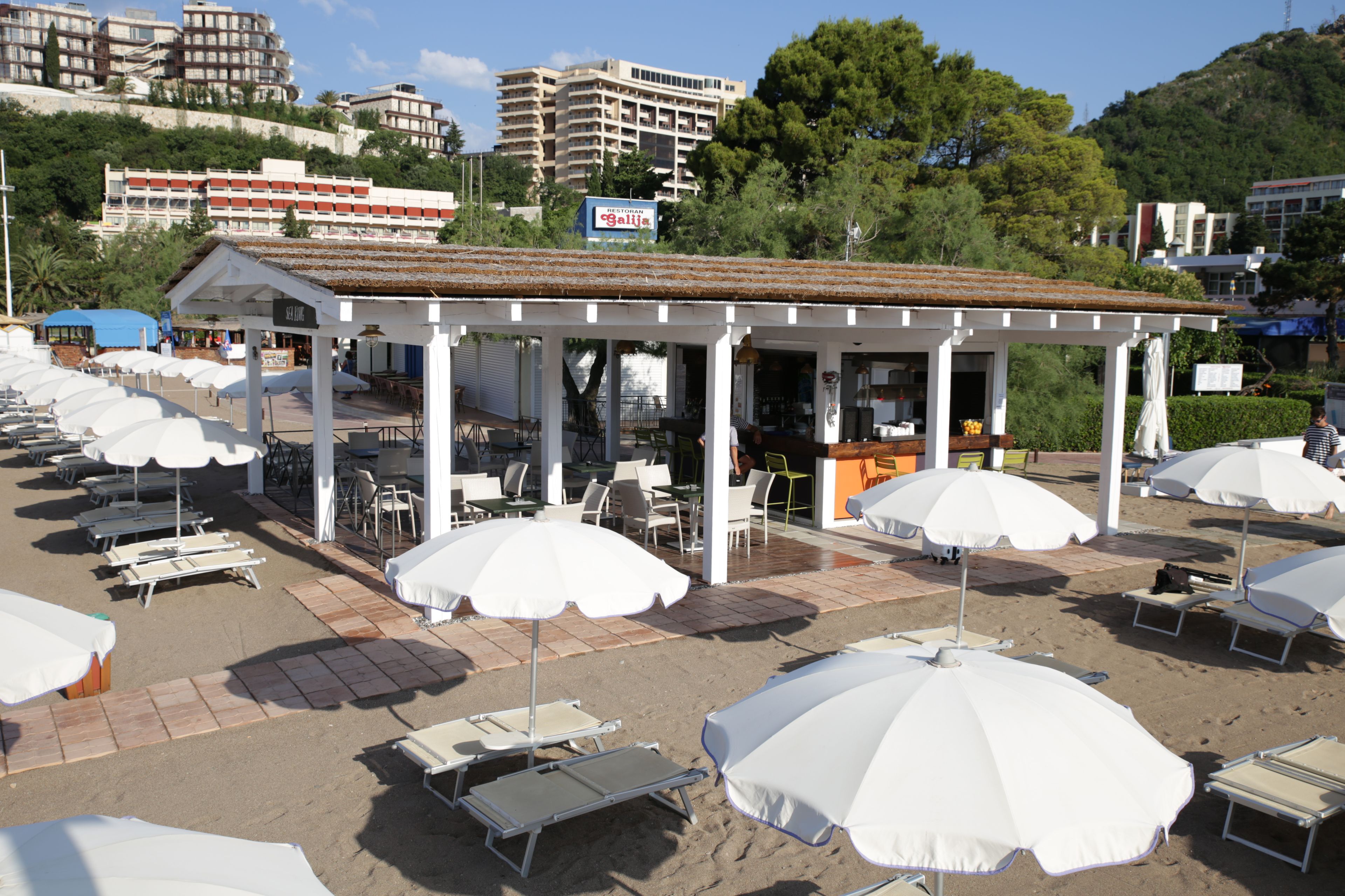 Un café situé en bord de mer, avec une terrasse, des parasols et une vue sur la ville en arrière-plan.