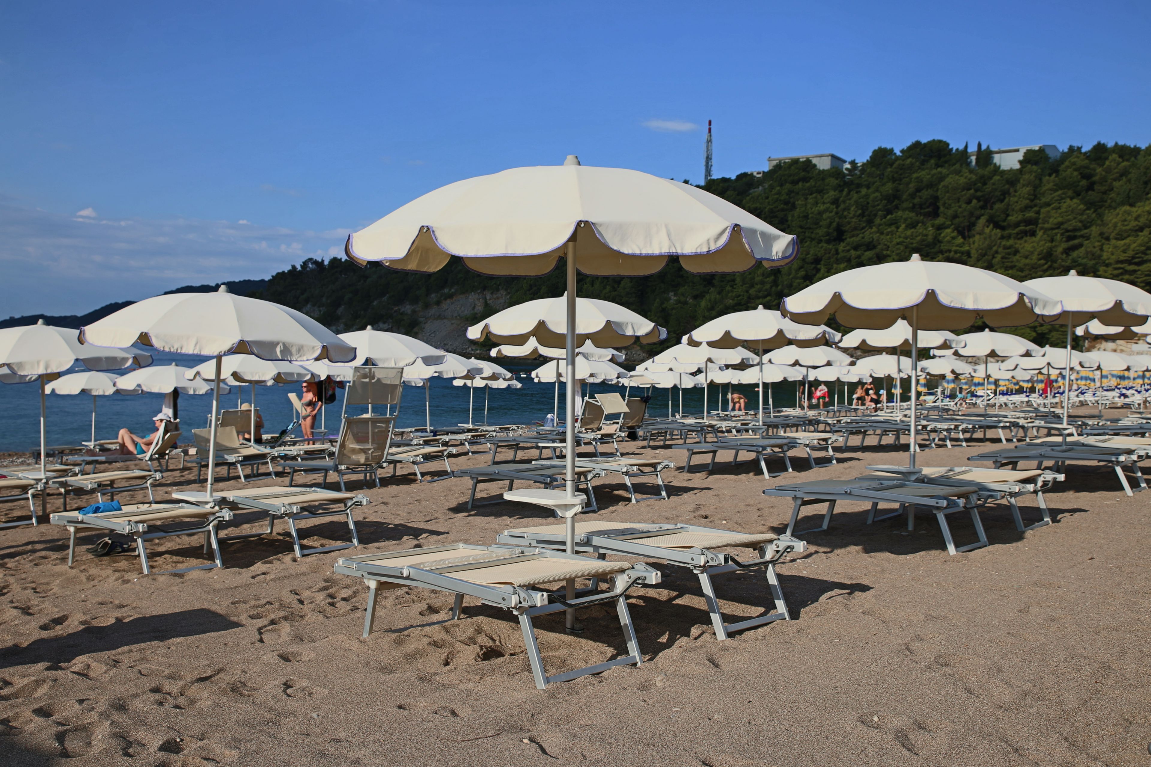 Une plage bordée de rangées de transats et de parasols, un ciel dégagé et une colline qui se dessine à l’horizon.