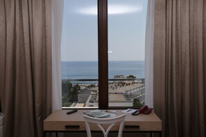 A room with a desk, a chair, a window with curtains, and a view of the sea and buildings. - Hotel Tara (Bečići)