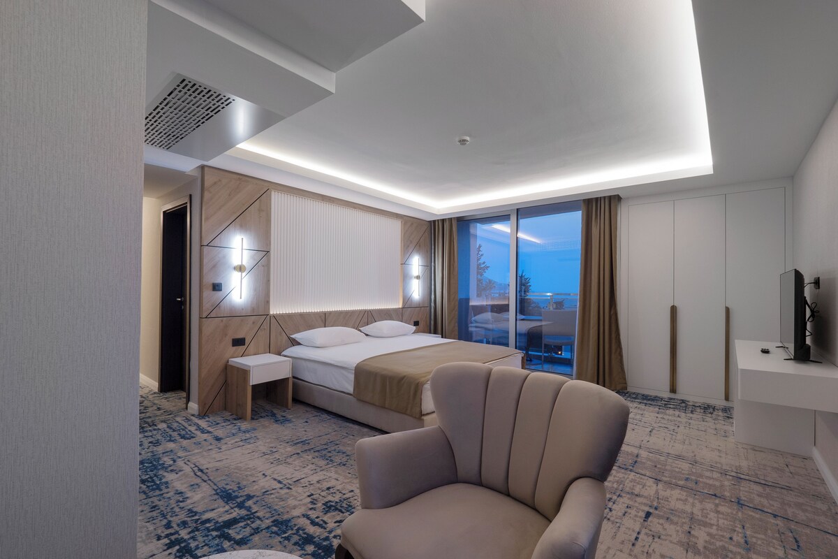 A modern hotel room with a bed, a chair, a desk, a TV, and a large window with a view.