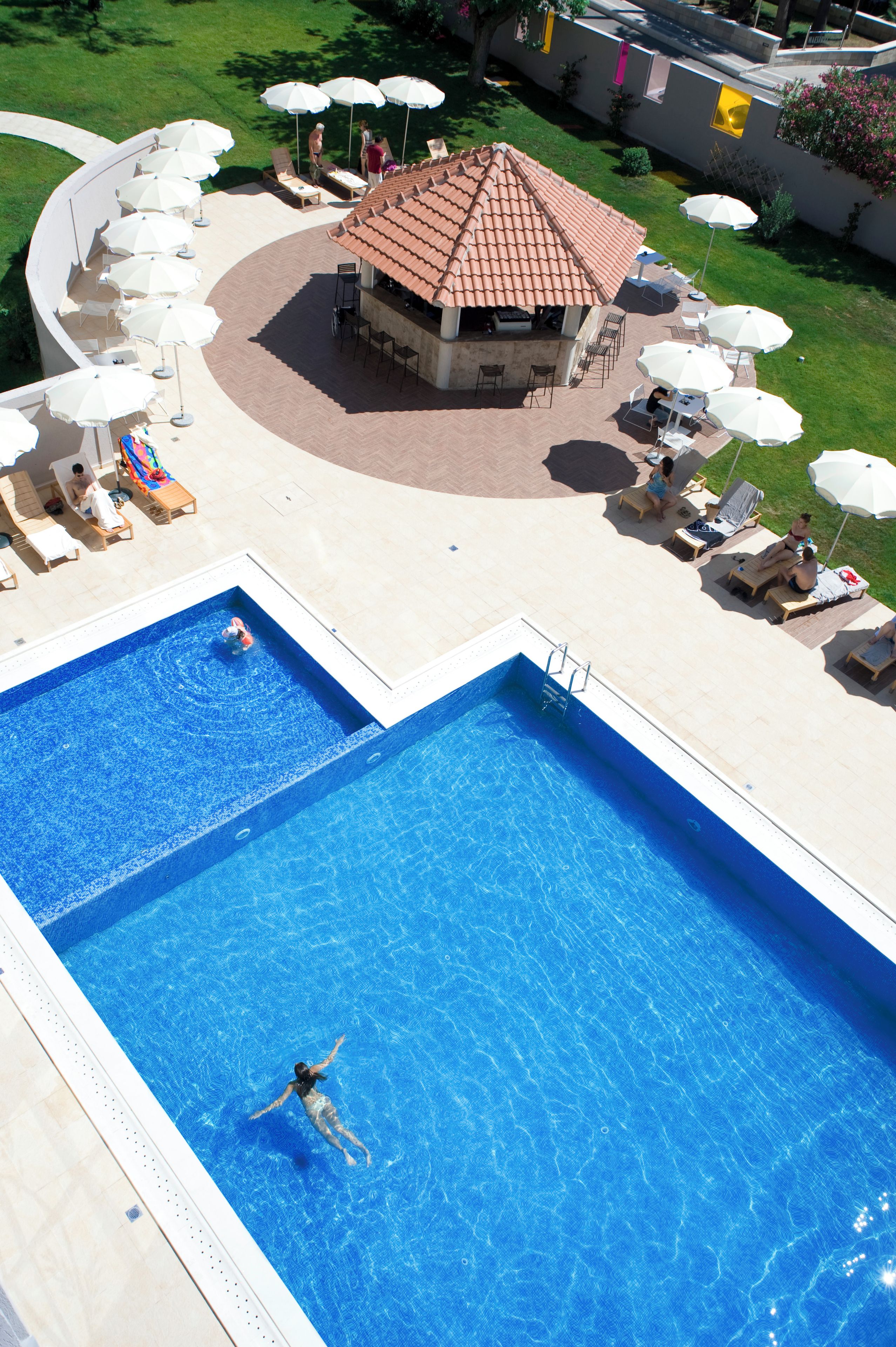 Una piscina con dos zonas diferenciadas, un patio cubierto con sillones y sombrillas, y un pequeño edificio con techo de tejas.