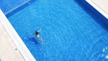 Una piscina con dos zonas diferenciadas, un patio cubierto con sillones y sombrillas, y un pequeño edificio con techo de tejas.