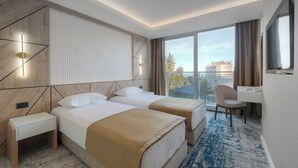 Habitación de hotel con dos camas, un escritorio, una silla, un televisor y un ventanal con vistas.