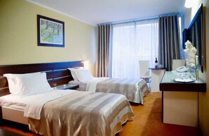 A hotel room with two beds, a desk, a chair, a window with curtains, and a painting on the wall. - Hotel Tara (Bečići)