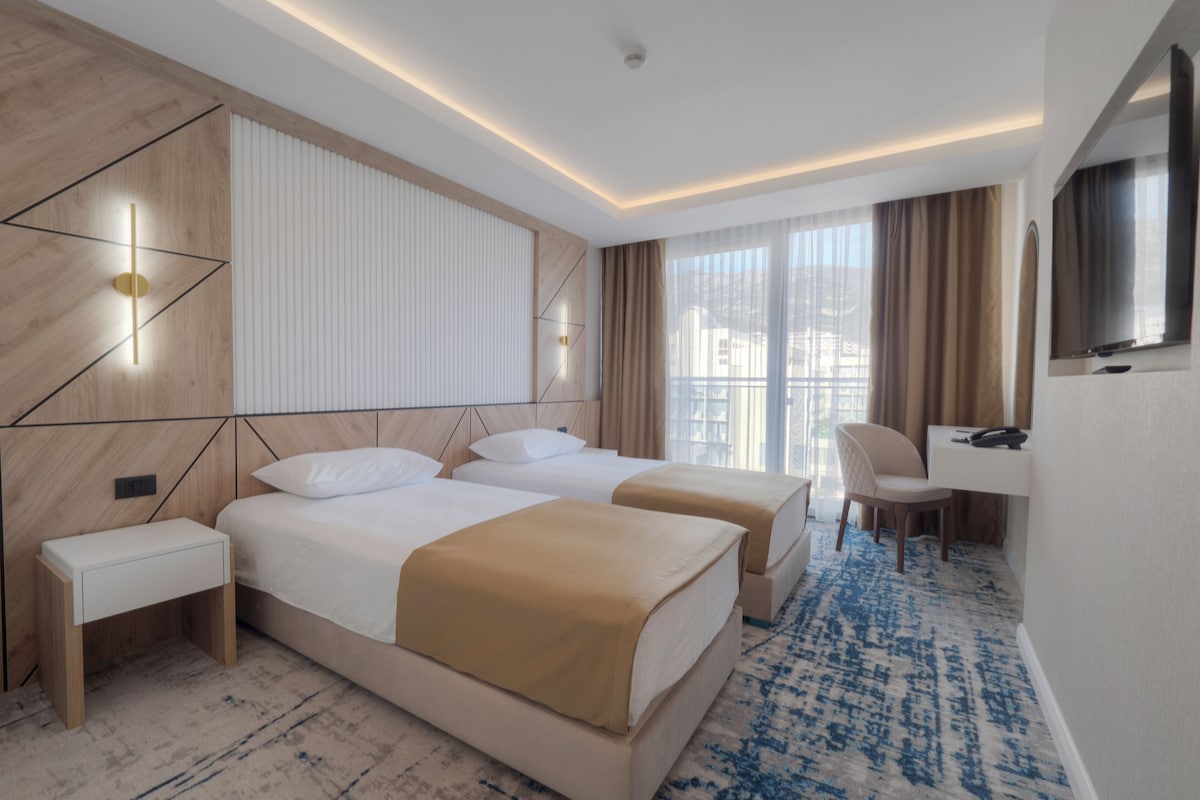 A hotel room with two beds, a desk, a chair, a TV, and a large window with curtains.