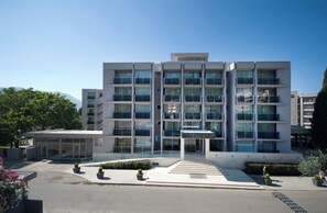 A multi-story hotel with a modern design, featuring a central entrance with steps, balconies on each floor, and a clear sky in the background. - Hotel Tara (Bečići)