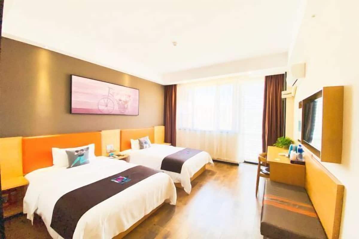 A hotel room with two beds, a desk, a chair, a TV, and a large window with curtains.