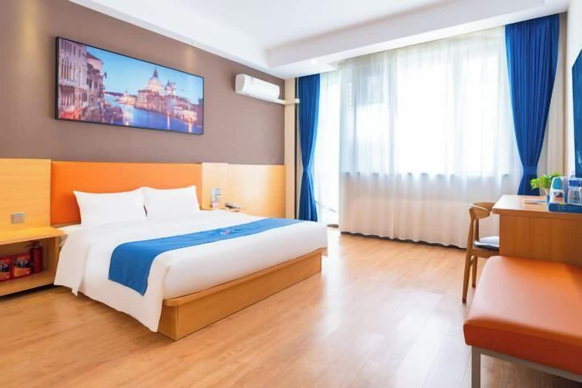 A hotel room with a bed, a desk, a chair, a painting, and a window with curtains.
