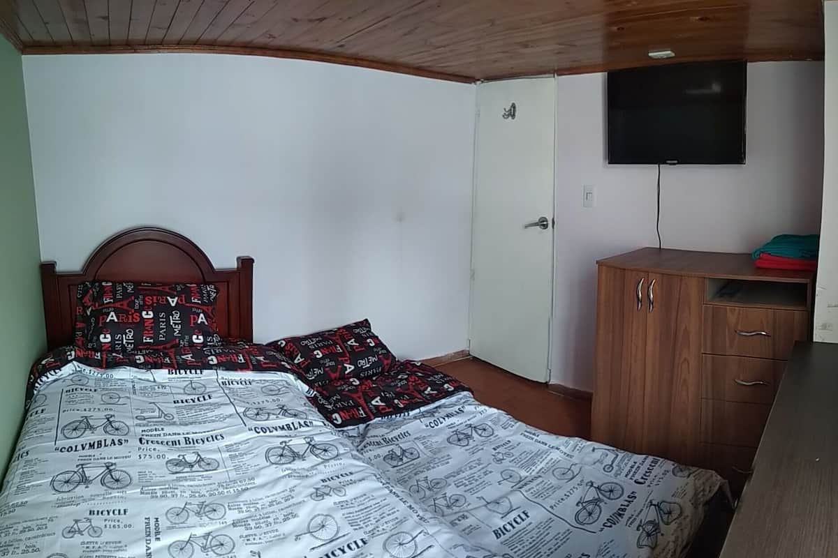 Ein Schlafzimmer mit einem hölzernen Kopfende, einem Bett mit einer Bettdecke mit Zeitungsmuster, einem Holzschrank, einem an der Wand montierten Fernseher und einer Tür.