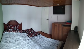 Une chambre avec une tête de lit en bois, un lit recouvert d’une couvre-lit à motifs de journaux, une armoire en bois, un téléviseur fixé au mur et une porte.