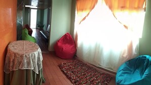 Ein Zimmer mit einem gemusterten Tisch, einem roten Sitzsack, einem farbenfrohen Teppich und Blick durch ein Fenster mit Vorhängen.