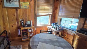 Una habitación de cabaña acogedora, con una cama, paredes de madera, un ventanal con persianas, una pequeña mesa con utensilios de cocina y un estante con diversos objetos.