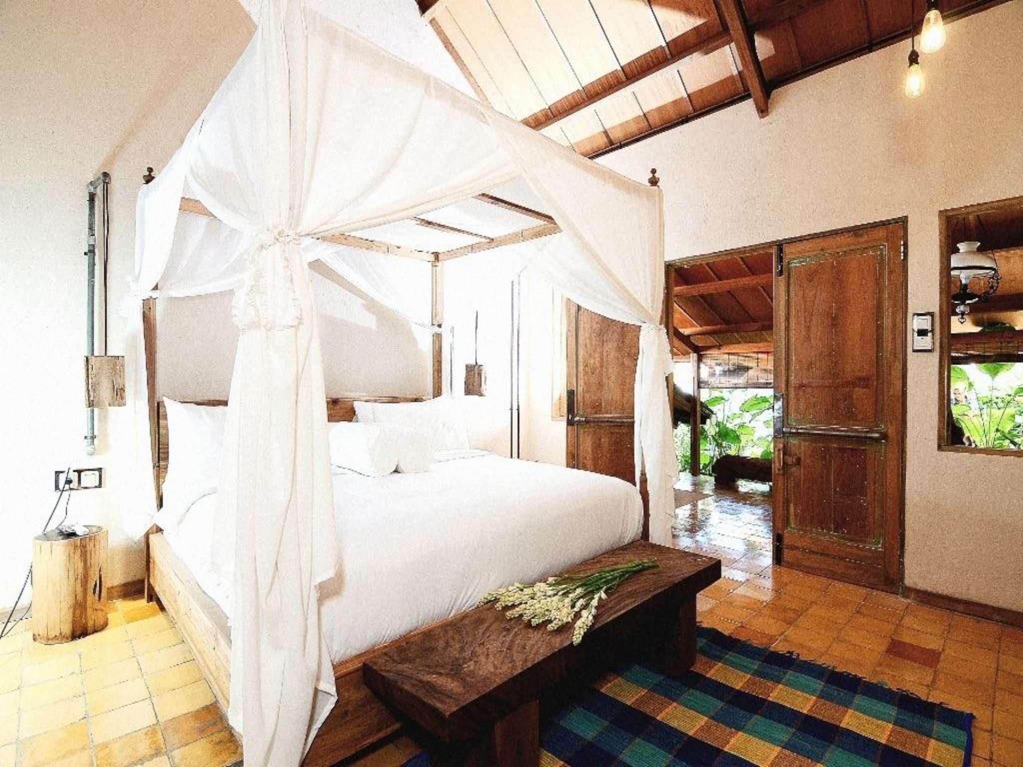 Een slaapkamer met een hemelbed, houten deuren, een nachtkastje met een bloem en een geruit vloertje.