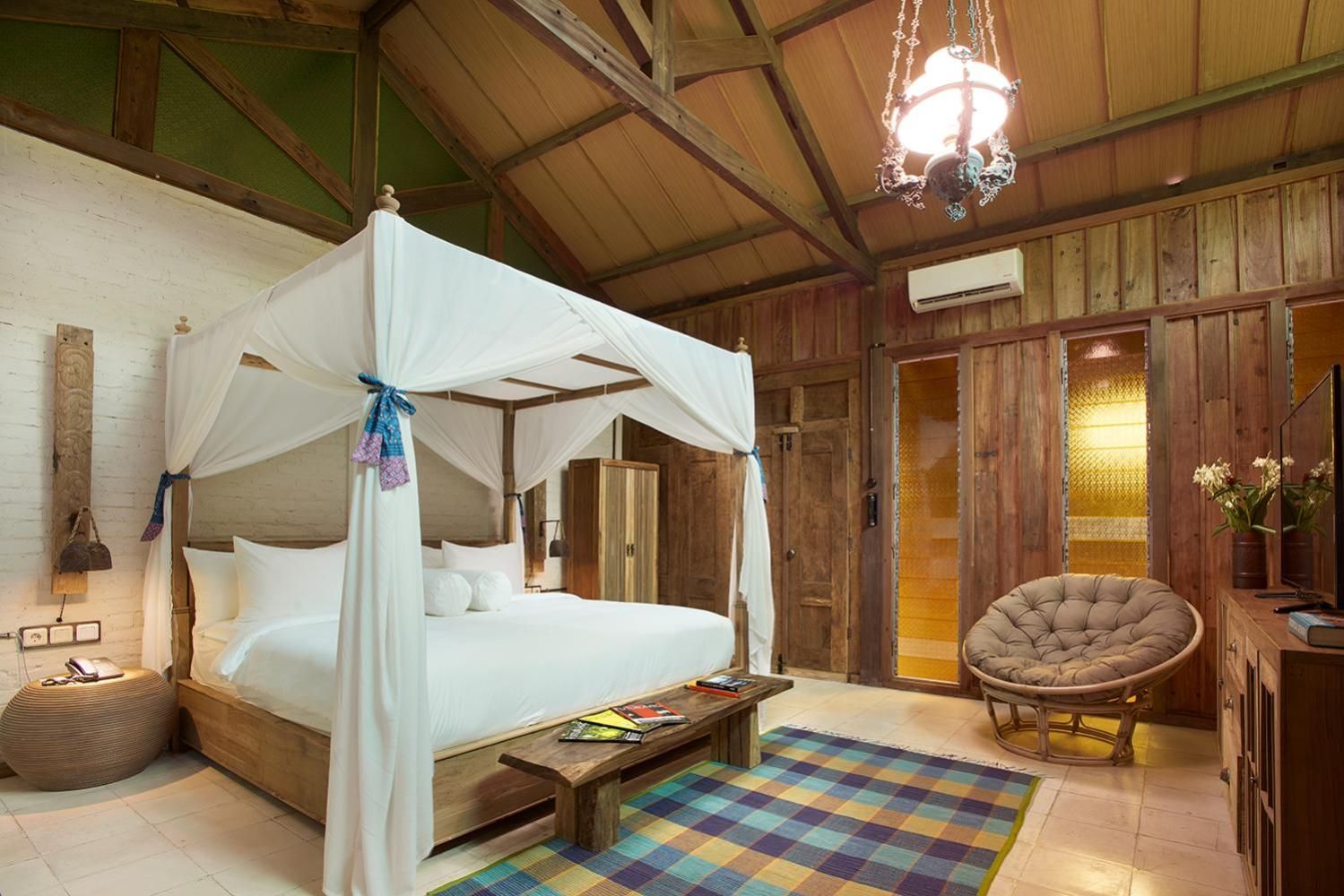 A four-poster canopy bed with a wooden headboard, a wooden bench, a colorful checkered rug, and a wicker chair.