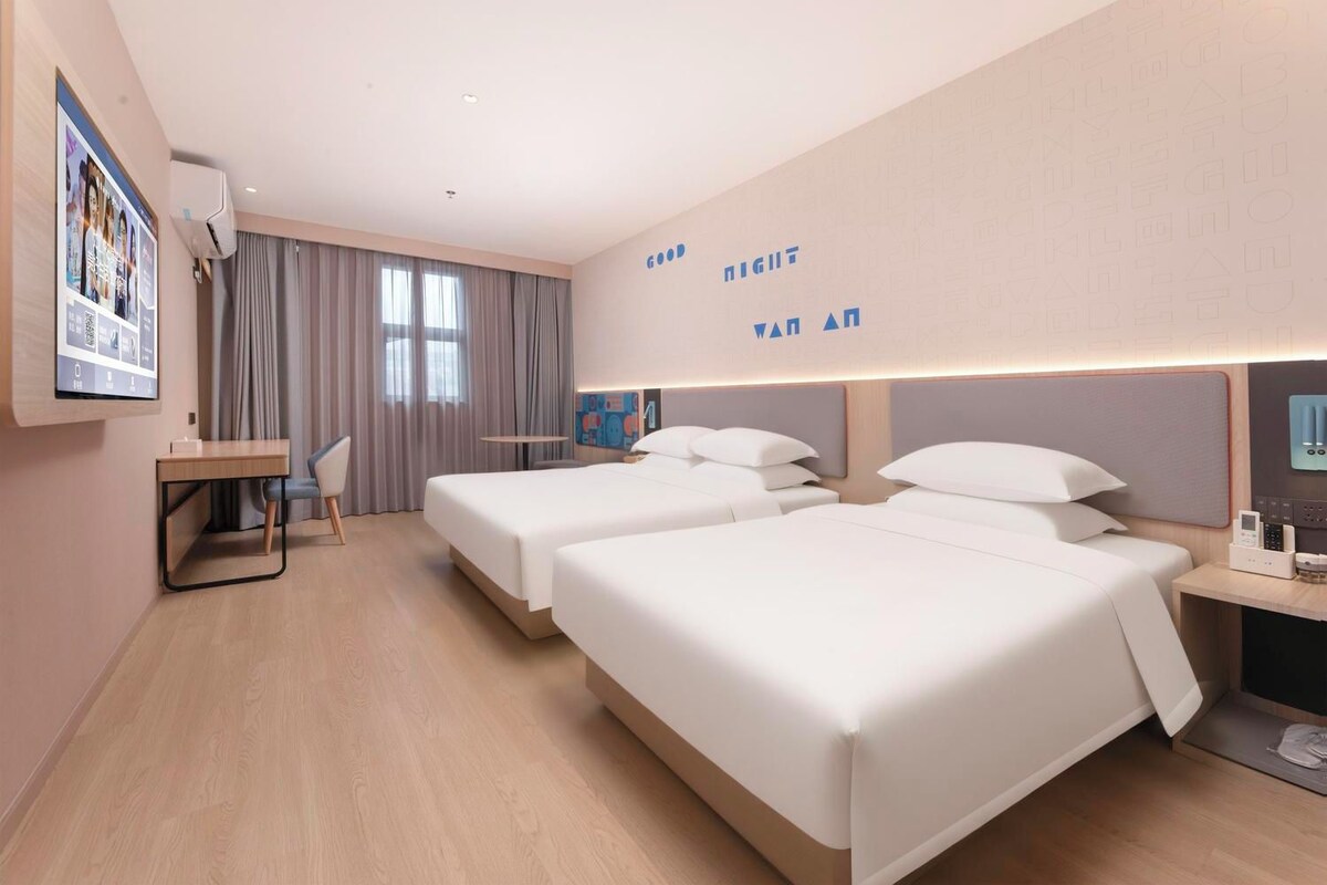 Habitación de hotel con dos camas, un escritorio, una silla y un amplio ventanal con cortinas.