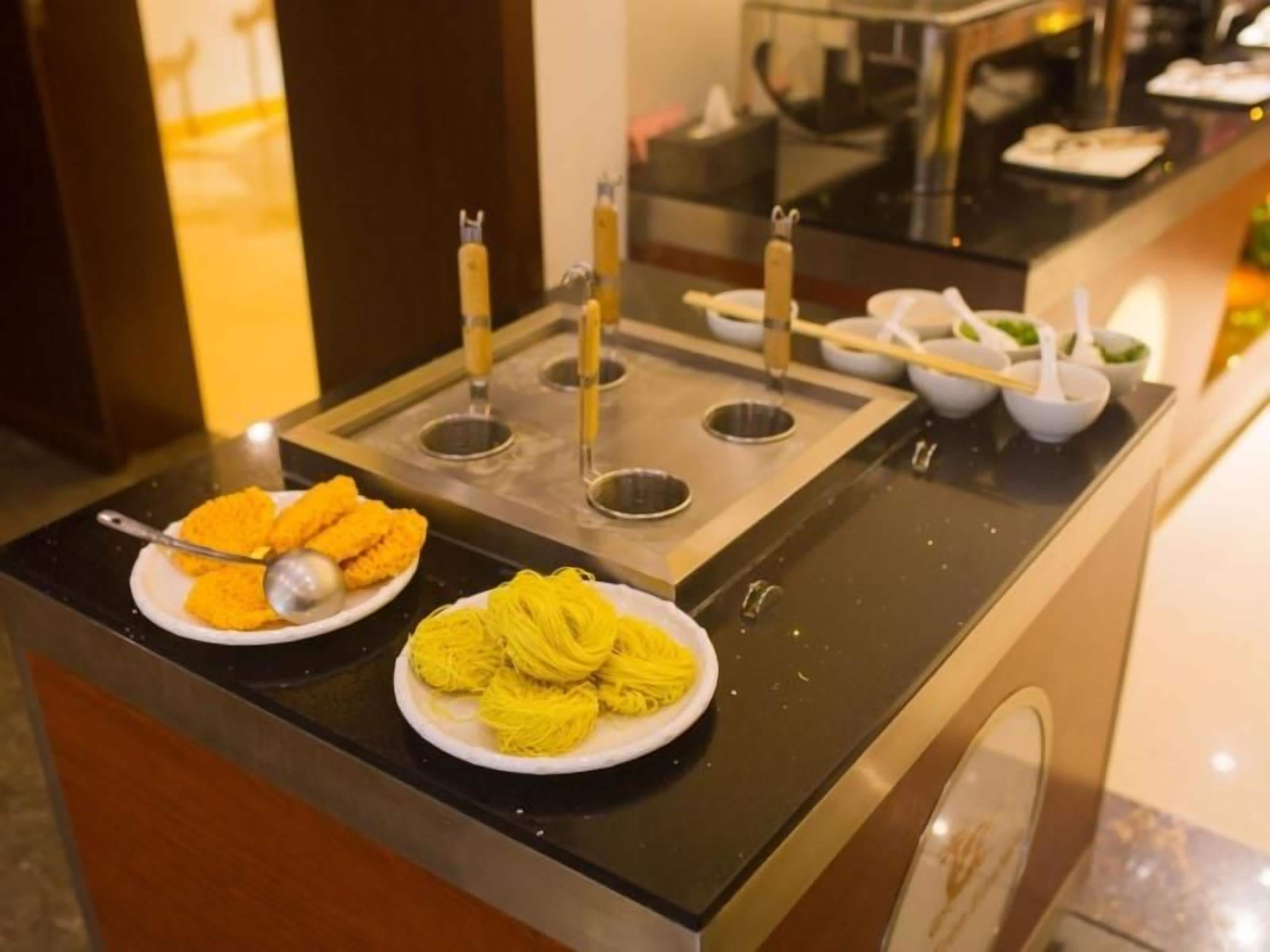 A buffet setup with two plates of food, one with orange-colored items and the other with yellow noodles, and a variety of utensils and bowls on a black countertop.