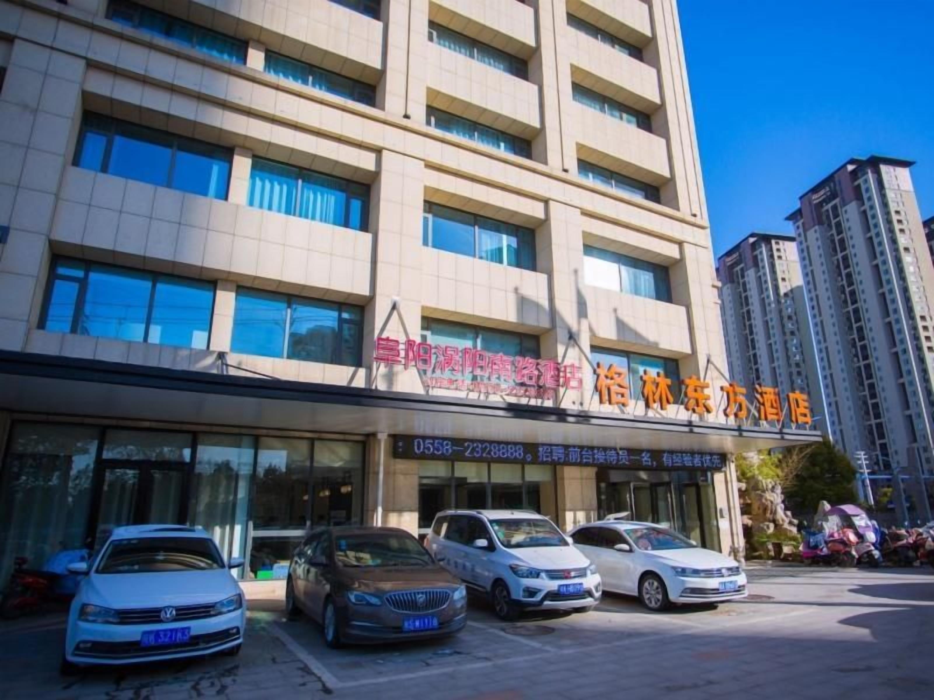 A multi-story hotel building with a clear sky. There are several cars parked in front, and the hotel's name is displayed on the facade.