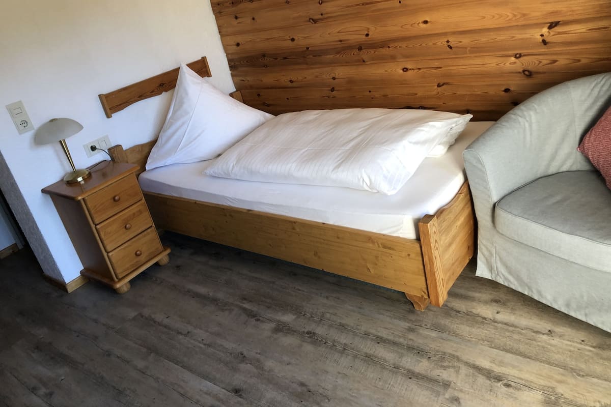 A wooden bed with white bedding, a wooden nightstand with a lamp, and a grey sofa.