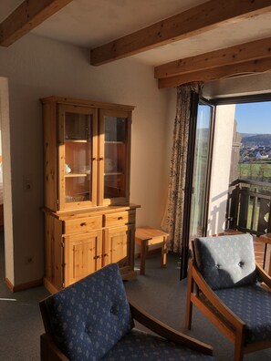 A room with a wooden cabinet, two blue patterned chairs, a small table, and a view of a town through a large window. - Landhotel Lortz (Reichelsheim)