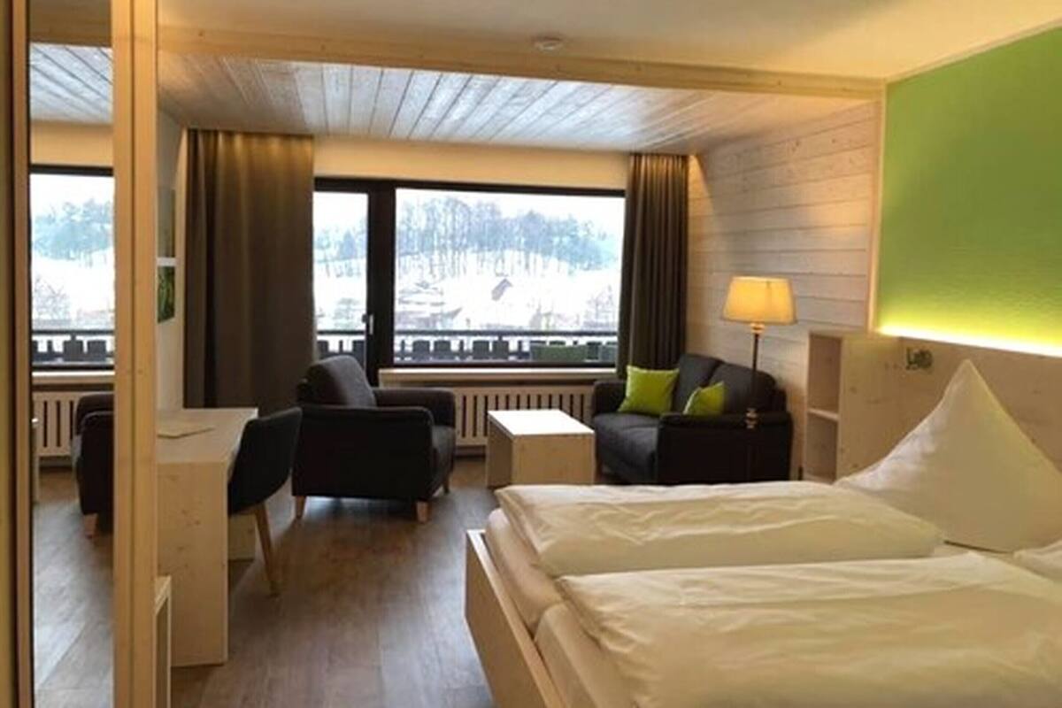 A hotel room with a bed, two chairs, a small table, a lamp, and a view of a snowy landscape through large windows.