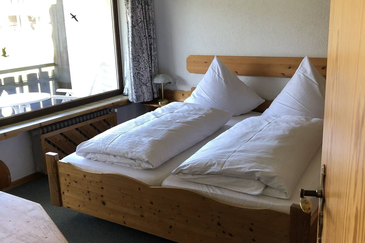 A wooden bed with white bedding, a window with curtains, and a wooden table.