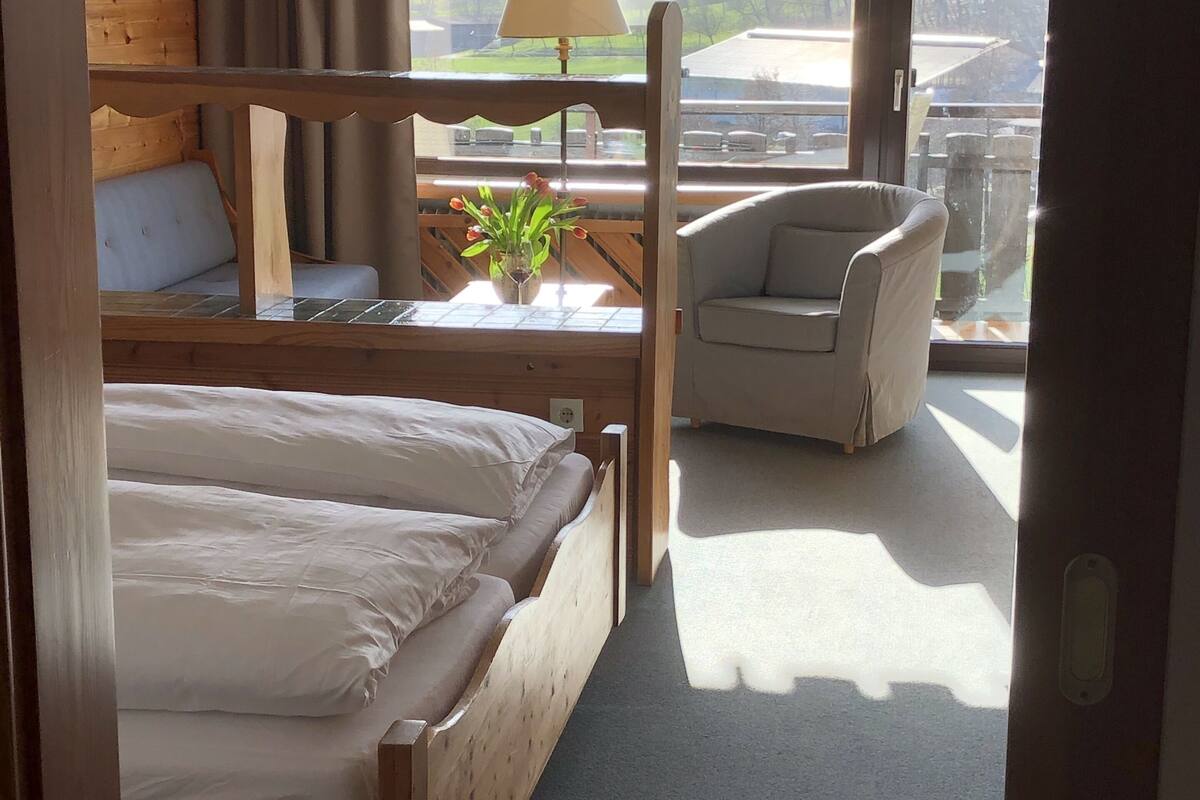 A bedroom with a wooden bed, a chair, a table with a lamp, and a view of the outdoors.