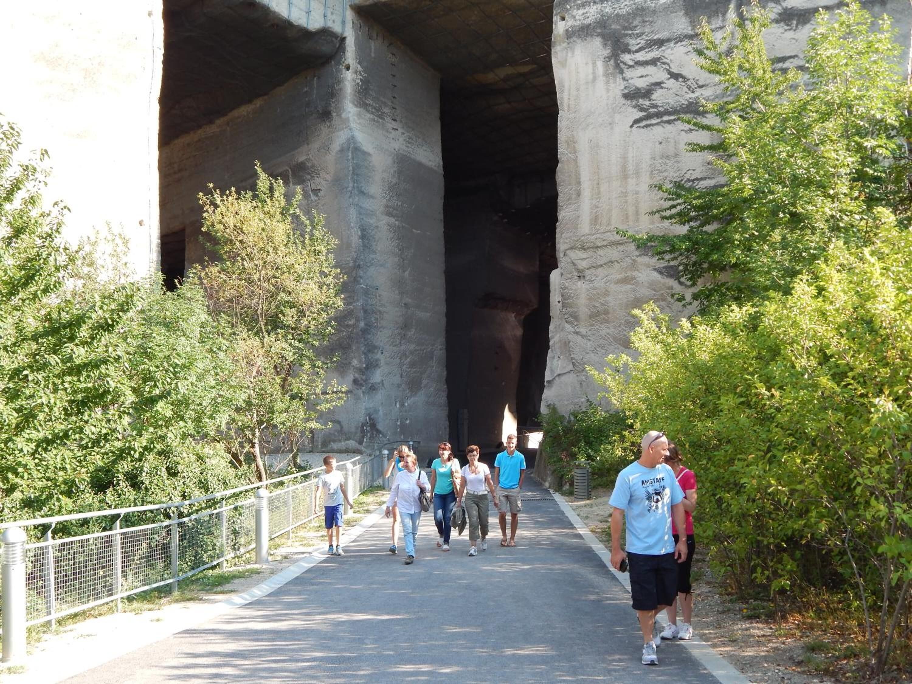 A pedestrian walkway under a large rock formation with a metal fence on one side and trees on the other.