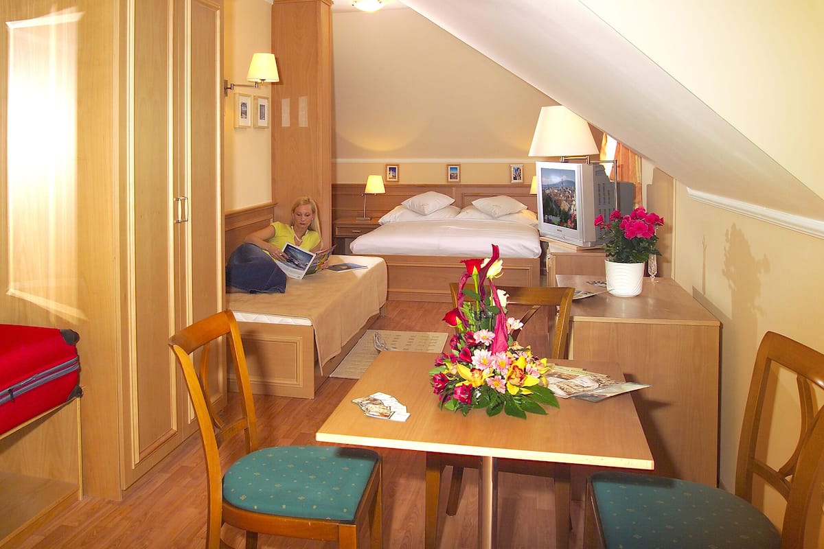 A compact hotel room with a bed, a small desk, a chair, and a table with a floral arrangement.