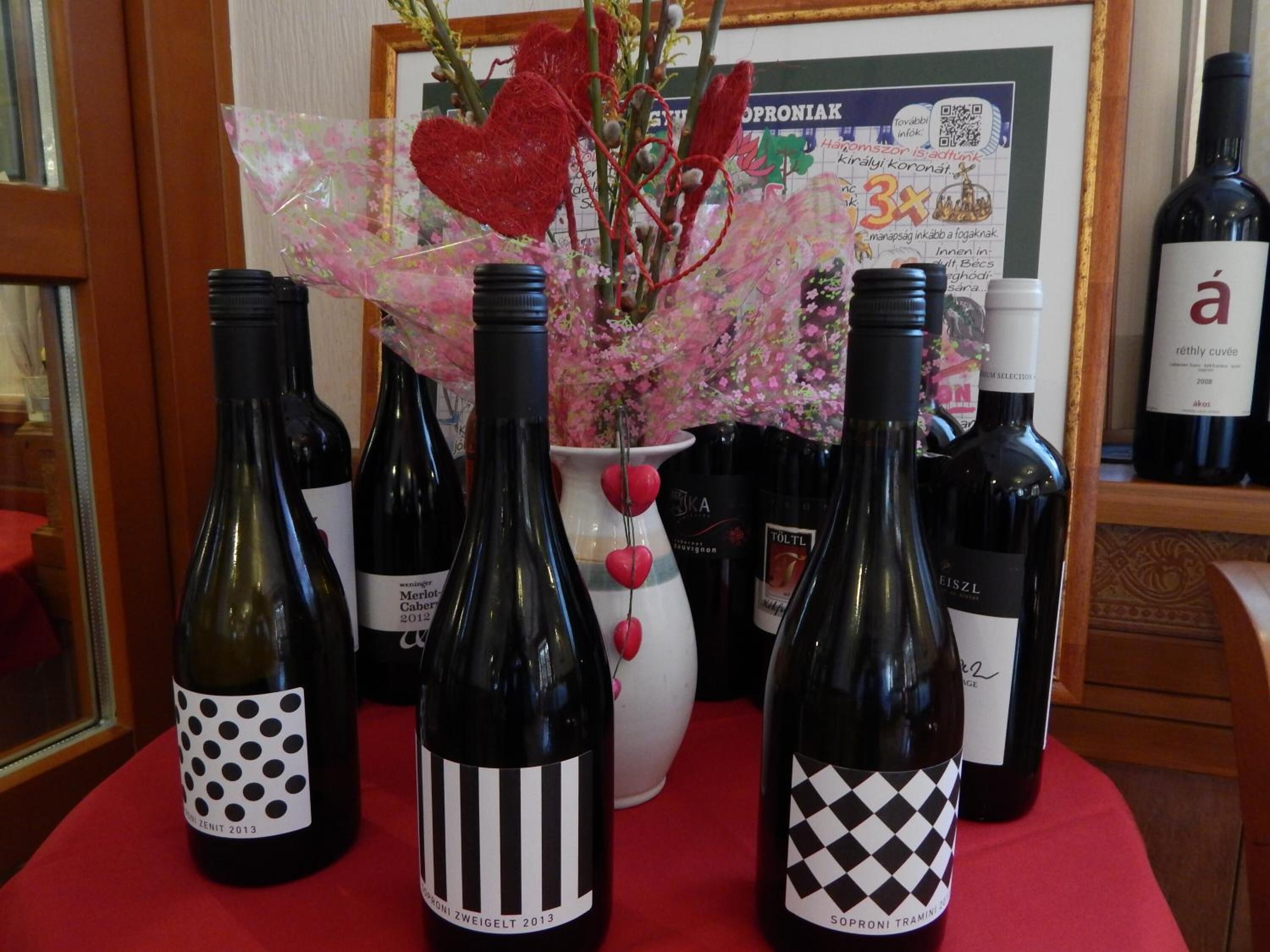 A selection of wine bottles on a table with a red cloth, a white vase with pink flowers, and a framed picture in the background.