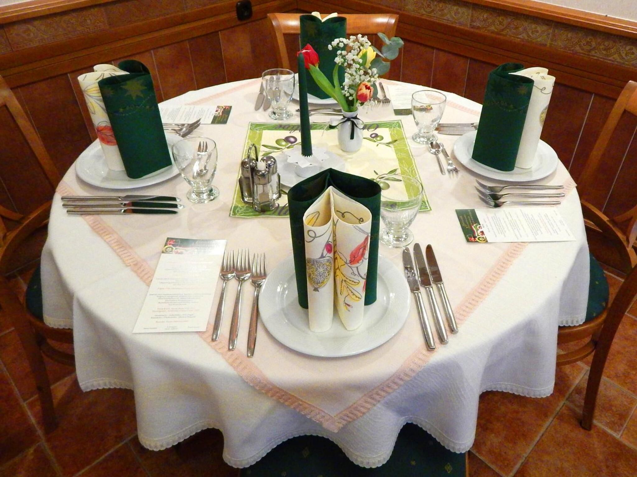Een ronde eettafel, gedekt met een witte tafelkleed, groene servetten en bestek.