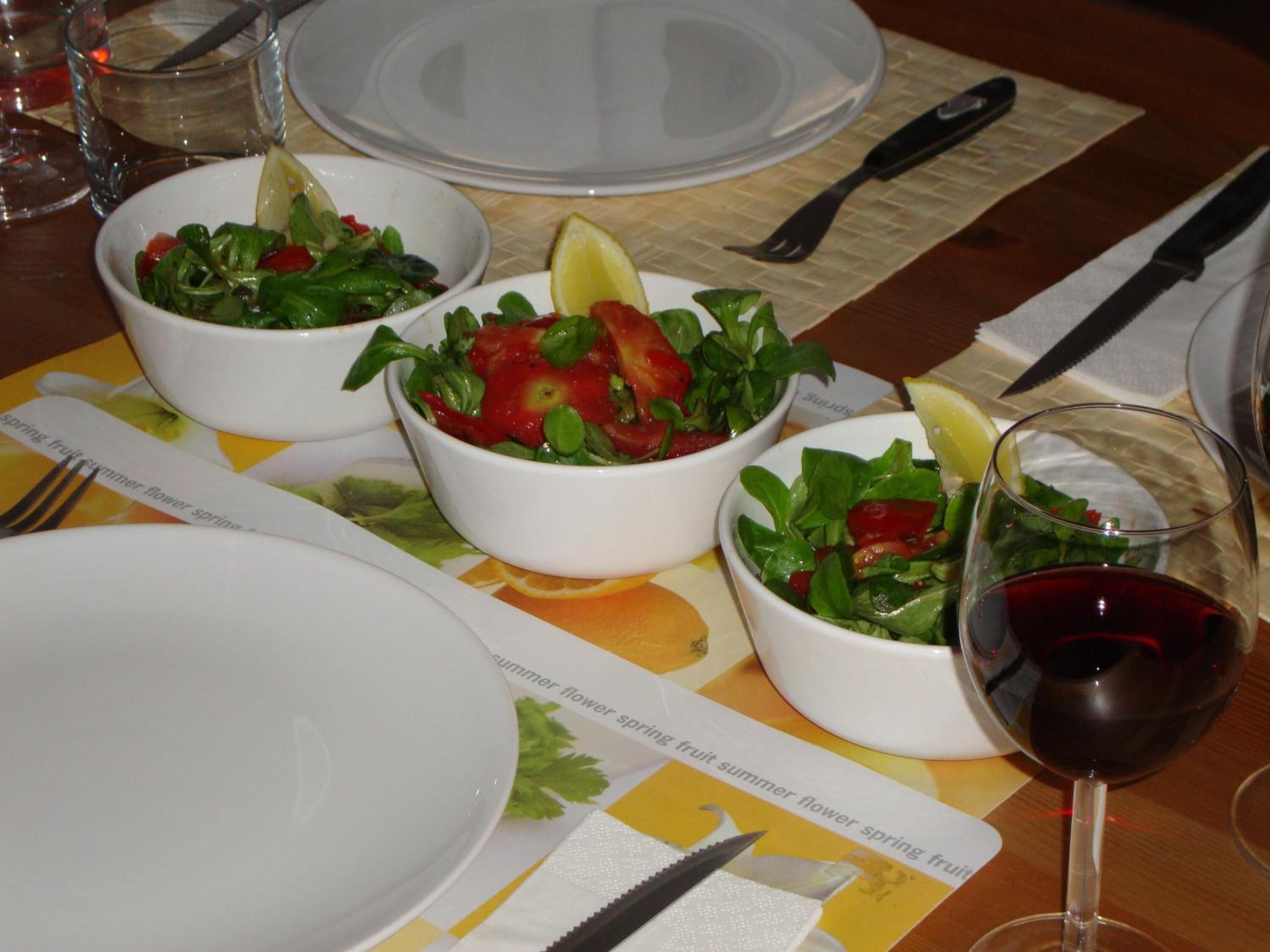 A table set with white bowls containing salads, a glass of red wine, and cutlery.