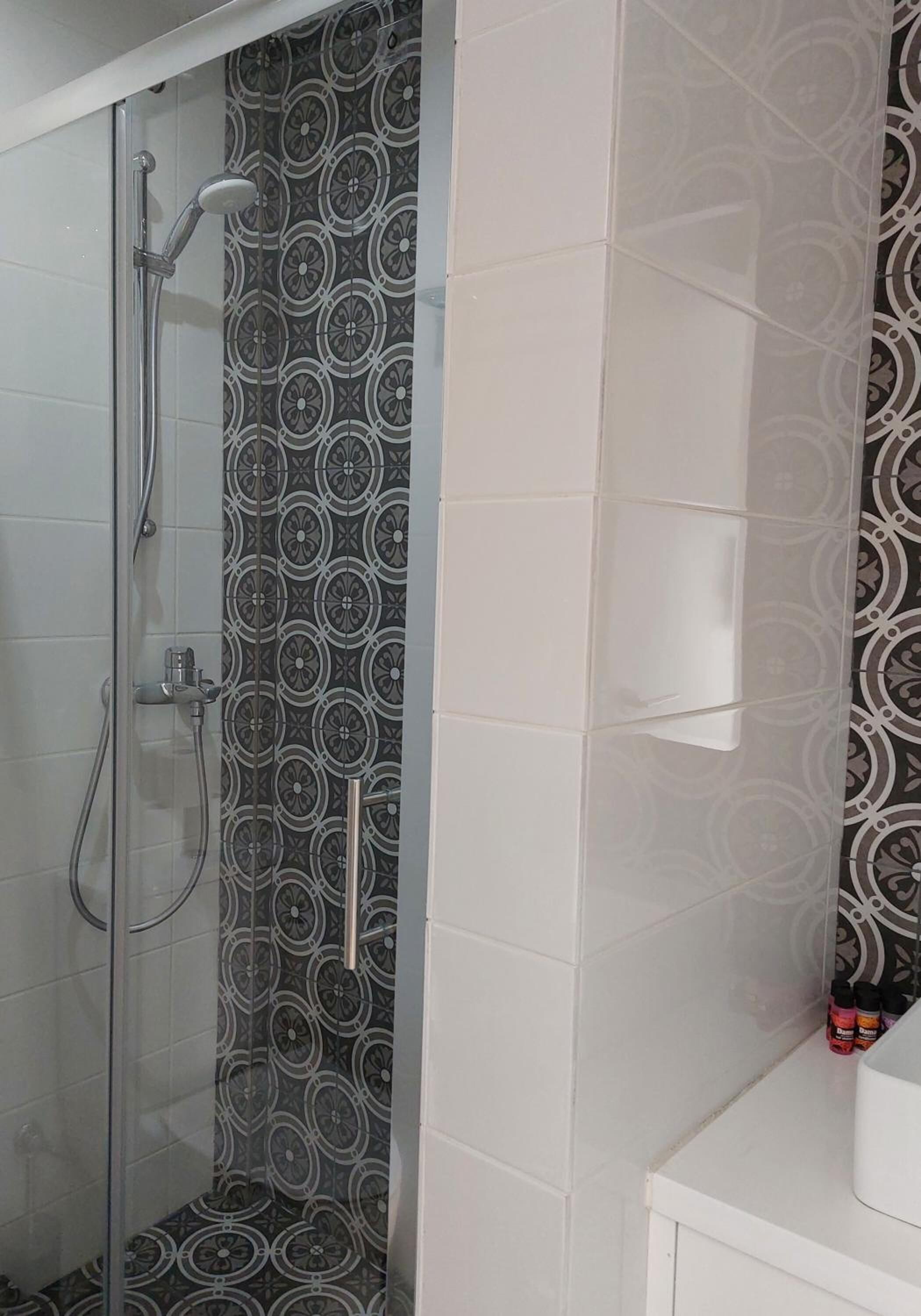 A shower with a glass door, a patterned wall, and a white tiled wall with a small shelf.