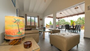 Un vaste espace de vie comprenant un canapé, un pouf et une table à manger. Une grande lampe au motif tropical est posée sur une table en bois.