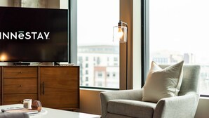Una habitación de hotel moderna con un televisor de pantalla plana grande, un sillón cómodo, una mesita de café y vistas a la ciudad a través de amplios ventanales.