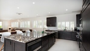 A modern kitchen with dark cabinetry, a large island with a granite countertop, and a dining area with white chairs. - Laguna Del Mar #08 (Seven Mile Beach)