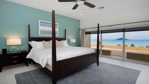 A bedroom with a four-poster bed, a nightstand with a lamp, a large window with a beach view, and a ceiling fan. - Laguna Del Mar #08 (Seven Mile Beach)