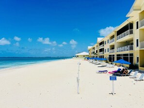 A beach with white sand, a clear blue sky, and a building with balconies and blue umbrellas.