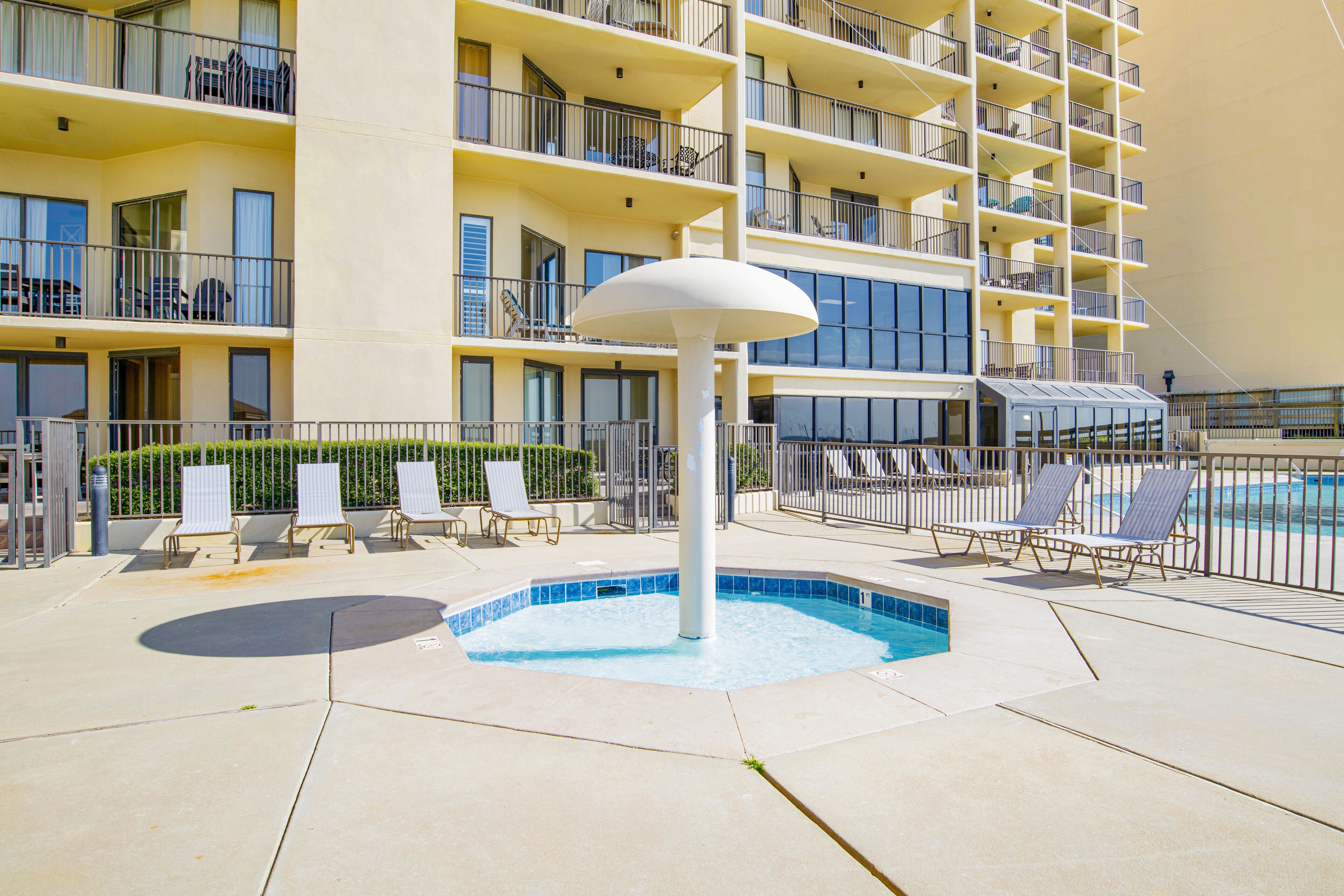 A hotel pool area with multiple balconies, lounge chairs, and a central water feature.