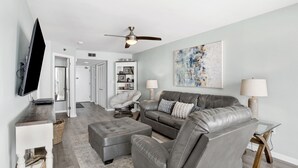A modern living room with a grey leather sofa, a matching ottoman, a flat-screen TV mounted on a wooden console, a ceiling fan, and a large abstract painting on the wall. - Beachfront 1BR @ Phoenix IV • Sleeps 4 • Stunning Views, Balcony & Resort Amenities (Orange Beach)