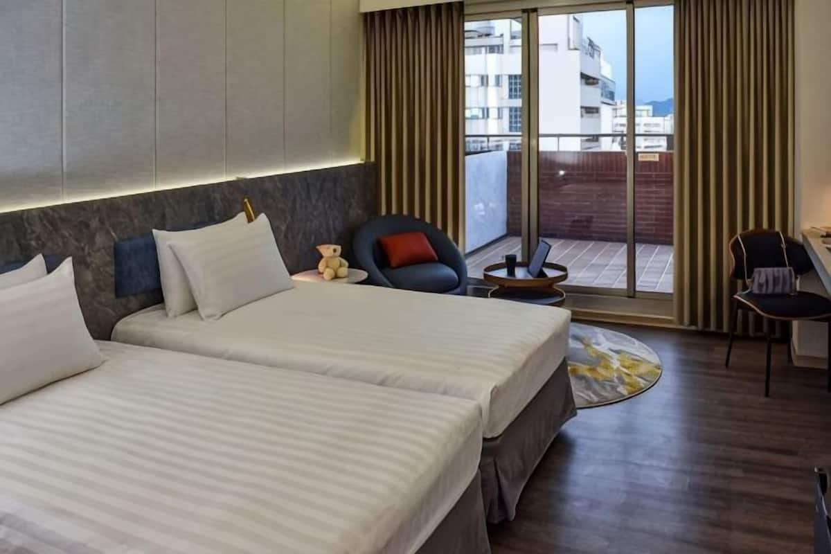 A hotel room with two beds, a desk, a chair, and a view of the outside.