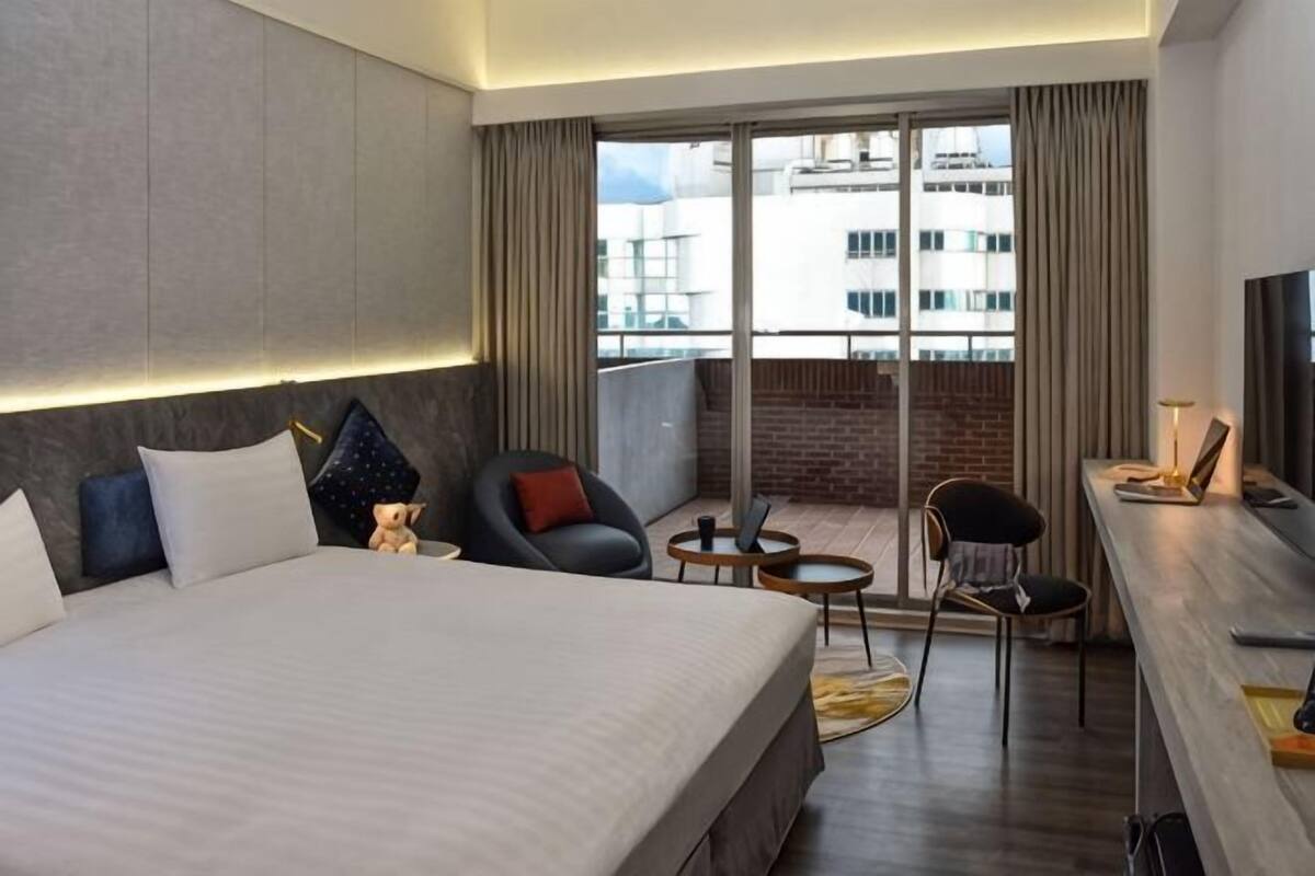 A modern hotel room with a large bed, a desk, a chair, and a balcony with a view of buildings.