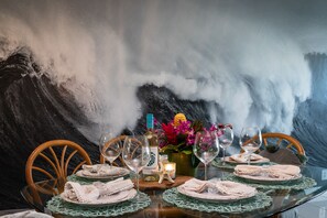 A dining table set with plates, glasses, and a bottle of wine, with a backdrop of a large wave.