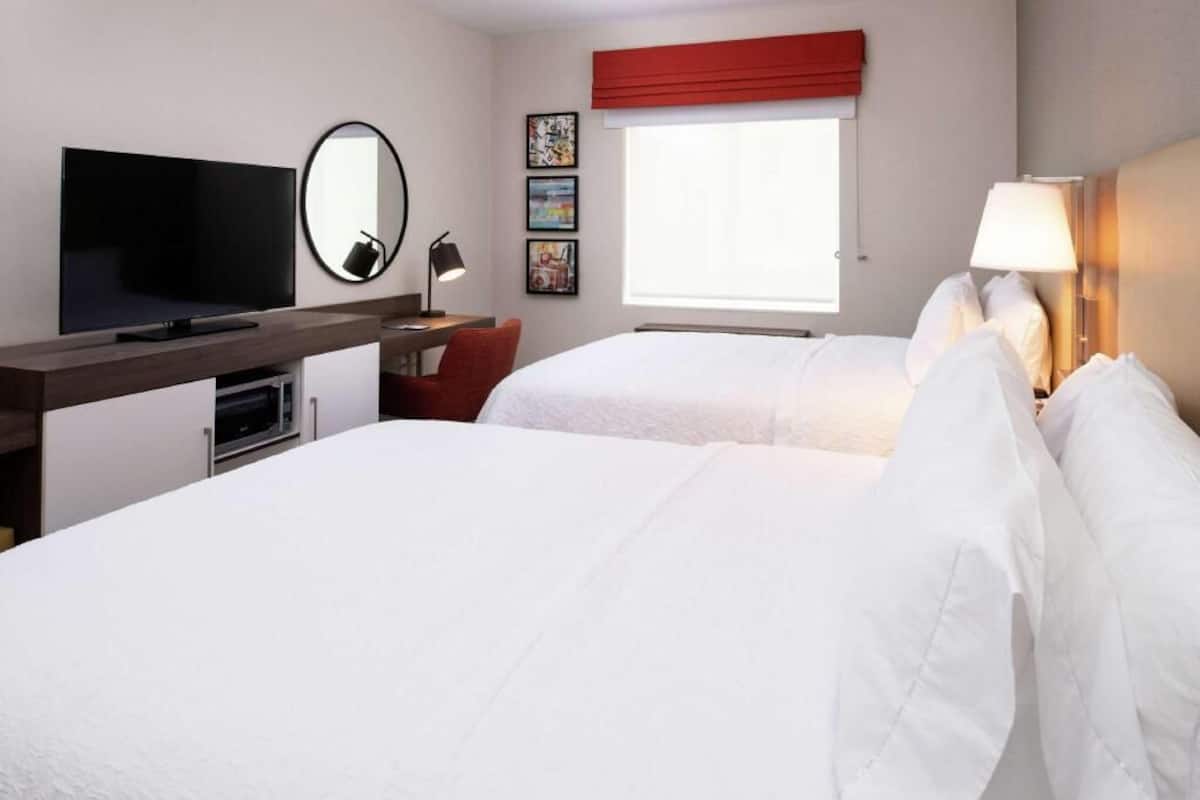 Une chambre d’hôtel équipée d’une télévision, d’un bureau avec une lampe, de deux lits, d’une fenêtre agrémentée d’un rideau rouge et d’œuvres d’art accrochées au mur.