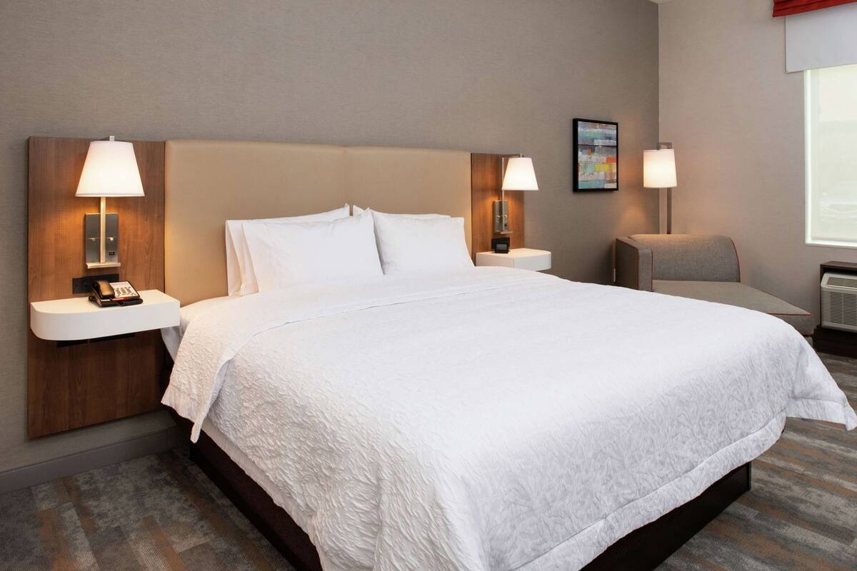 Une chambre d’hôtel avec un grand lit, deux tables de chevet avec des lampes, une chaise et une fenêtre avec des stores.