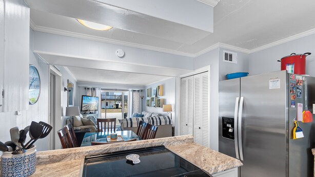 A modern kitchen with a built-in stove, stainless steel refrigerator, and a dining area with a glass table and chairs. - "Shore Beats Work" - 2nd Row Condo w/ Community Pool, 4 Bedrooms / 2 Bathrooms (Holden Beach)