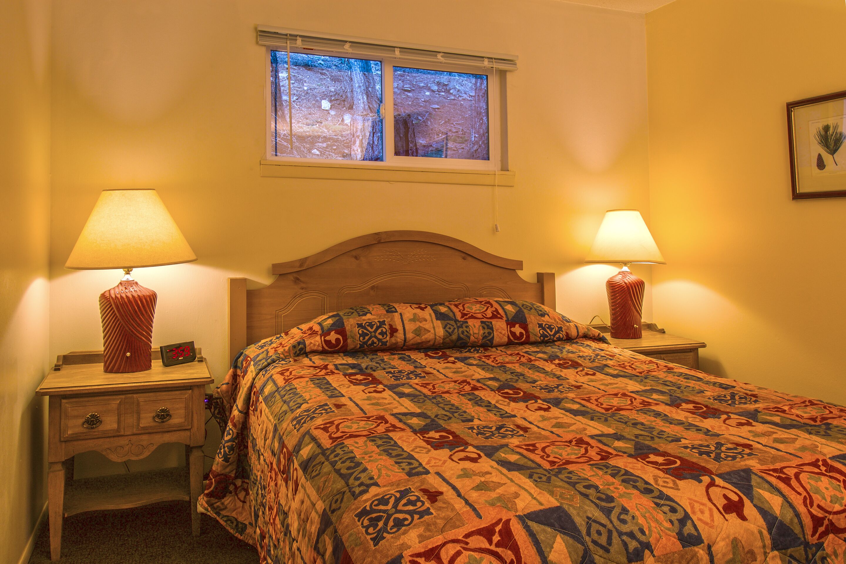 A bedroom with a bed, two bedside lamps, a nightstand with a clock, and a window with a view of trees.