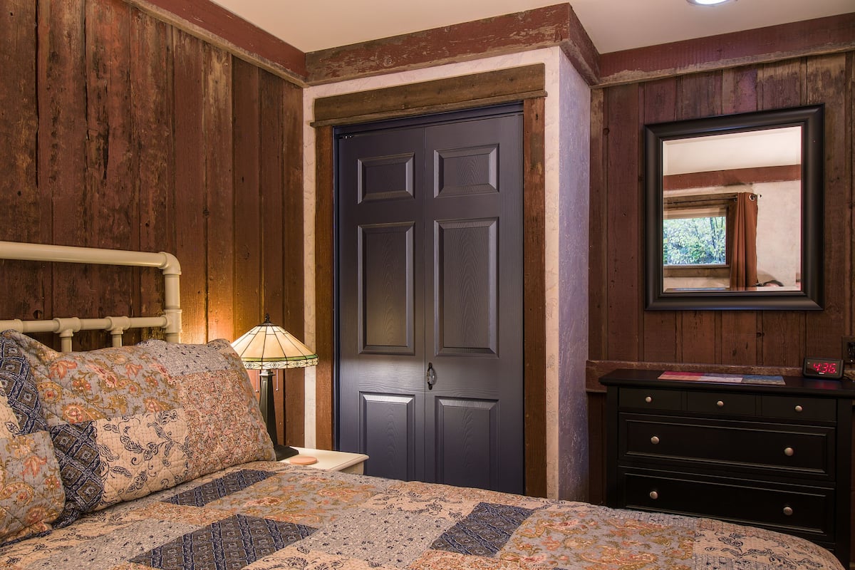 Un dormitorio con pared de madera, una puerta oscura, una cama con ropa de cama estampada, una mesita de noche con lámpara y un aparador con espejo.