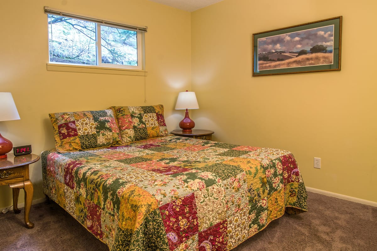 Un dormitorio con una colcha floral acolchada, una mesita de noche con una lámpara, un reloj y una ventana con vistas a árboles.