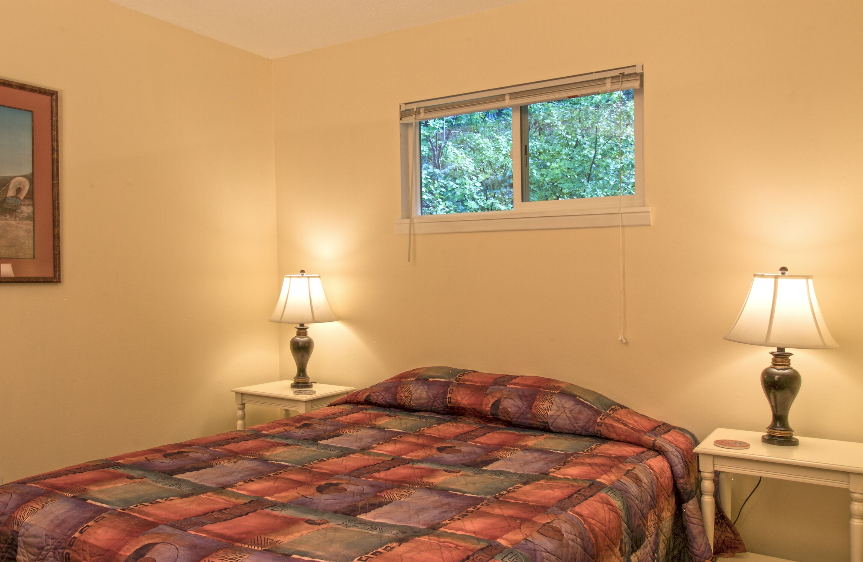 Un dormitorio con cama, dos mesitas de noche con lámparas, una ventana con vistas a árboles y un cuadro en la pared.