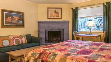 Une pièce chaleureuse avec une cheminée, un lit recouvert d’une couverture aux couleurs vives, un canapé agrémenté d’un plaid à motifs et un tableau accroché au mur.